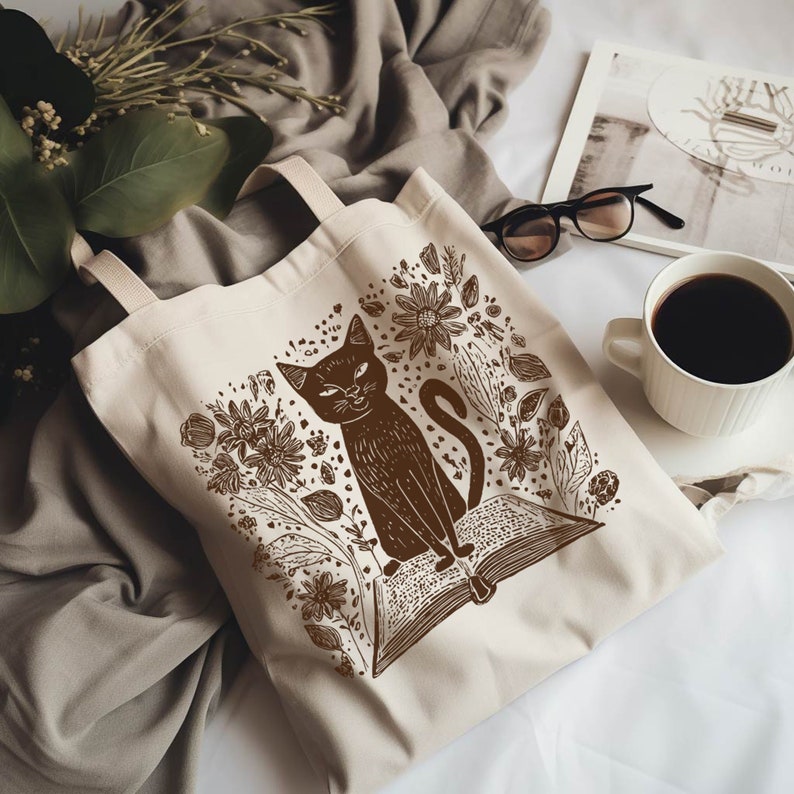 May include: A brown tote bag with a black cat sitting on an open book surrounded by flowers. The cat is looking to the right. The bag is made of canvas and has a brown handle.