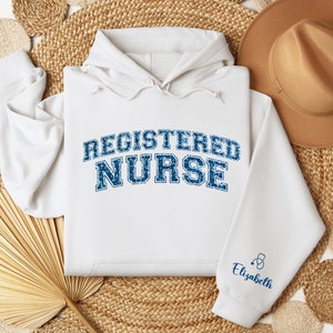 May include: White hoodie with the text "REGISTERED NURSE" in blue with a distressed look. The sleeve has a small blue stethoscope and the name "Elizabeth" in blue text.