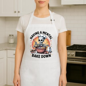 May include: White apron featuring the text "HAVING A MENTAL BAKE DOWN" and a cartoon skeleton chef. The skeleton is wearing a chef's hat and mixing ingredients in a bowl with a rolling pin and eggs.