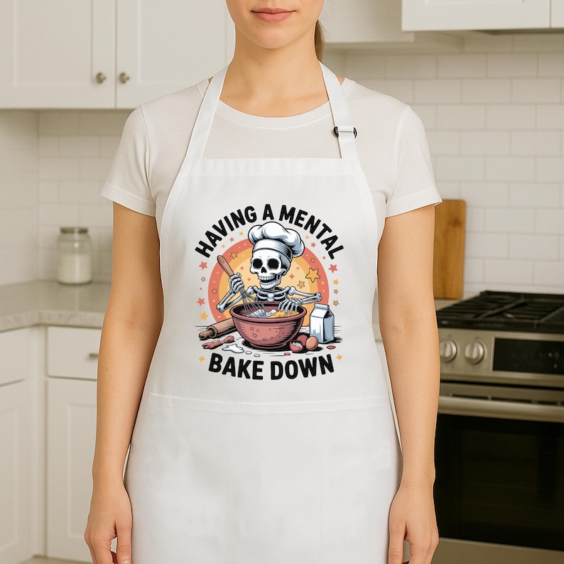 May include: White apron featuring the text "HAVING A MENTAL BAKE DOWN" and a cartoon skeleton chef. The skeleton is wearing a chef's hat and mixing ingredients in a bowl with a rolling pin and eggs.