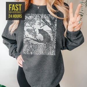 Peut inclure: Sweat-shirt gris fonc&eacute; avec un motif noir et blanc d'un chat perch&eacute; sur une branche d'arbre. Le motif comprend une illustration d&eacute;taill&eacute;e d'un arbre, de fleurs et de hautes herbes. Le sweat-shirt porte &eacute;galement le texte "FAST SHIPPING 24 HOURS" dans un encadr&eacute; jaune.