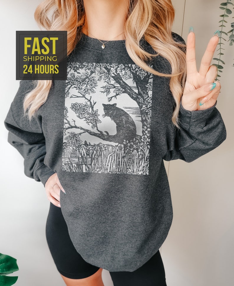 Peut inclure: Sweat-shirt gris fonc&eacute; avec un motif noir et blanc d'un chat perch&eacute; sur une branche d'arbre. Le motif comprend une illustration d&eacute;taill&eacute;e d'un arbre, de fleurs et de hautes herbes. Le sweat-shirt porte &eacute;galement le texte "FAST SHIPPING 24 HOURS" dans un encadr&eacute; jaune.