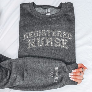 May include: Gray sweatshirt with the text "REGISTERED NURSE" in silver glitter. The sweatshirt has a small embroidered stethoscope and the name "Elizabeth" on the sleeve.