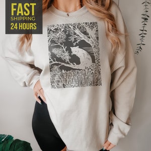 Peut inclure: Un sweat-shirt beige clair avec un motif noir et blanc d'un chat dans une for&ecirc;t. Le sweat-shirt a des manches longues et un col rond. Le texte "FAST SHIPPING 24 HOURS" est dans un encadr&eacute; jaune en haut &agrave; gauche.