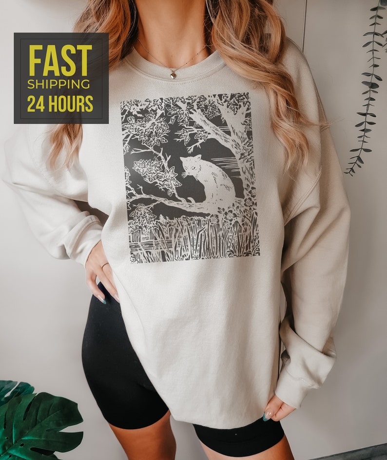 Peut inclure: Un sweat-shirt beige clair avec un motif noir et blanc d'un chat dans une for&ecirc;t. Le sweat-shirt a des manches longues et un col rond. Le texte "FAST SHIPPING 24 HOURS" est dans un encadr&eacute; jaune en haut &agrave; gauche.