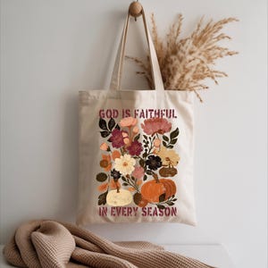 A beige tote bag with a floral design and the text "God is Faithful in Every Season" printed on it, hanging on a hook against a white wall.