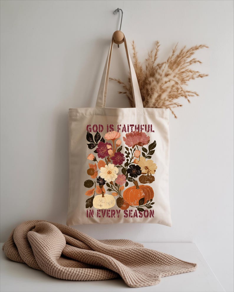 A beige tote bag with a floral design and the text "God is Faithful in Every Season" printed on it, hanging on a hook against a white wall.