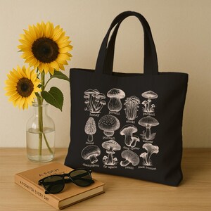 May include: Black canvas tote bag with a white illustration of different types of mushrooms. The mushrooms are labeled with their names, including honey, porcini, enoki, parasol, morel, puffball, king oyster, weboap, champignon, fly agaric, reishi, and black trumpet.