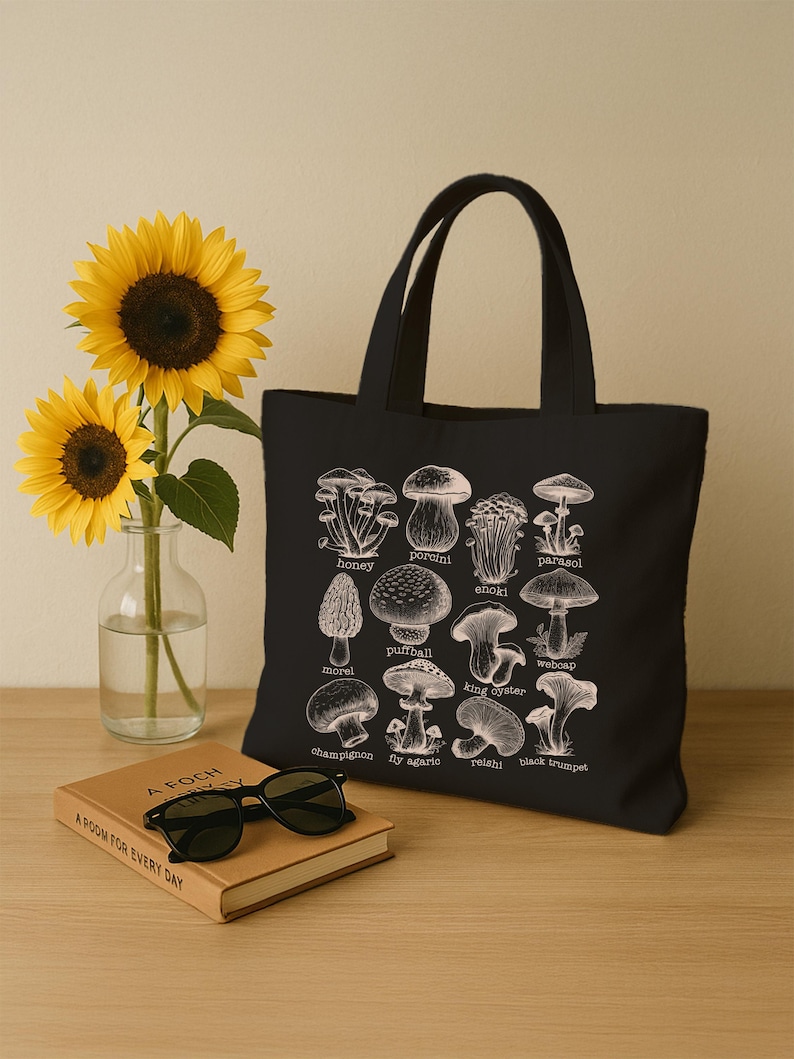May include: Black canvas tote bag with a white illustration of different types of mushrooms. The mushrooms are labeled with their names, including honey, porcini, enoki, parasol, morel, puffball, king oyster, weboap, champignon, fly agaric, reishi, and black trumpet.