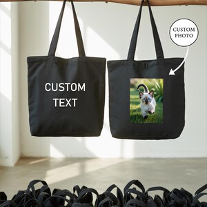 two black tote bags hanging from a wooden bar, with one bag having a custom photo printed on it.