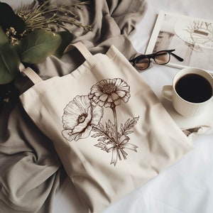 K&ouml;nnte beinhalten: Eine wei&szlig;e Canvas-Tragetasche mit braunen floralen Illustrationen von zwei Blumen, eine mit einer gro&szlig;en Mitte und die andere mit einer kleineren Mitte. Die Blumen sind mit einem Band zusammengebunden.