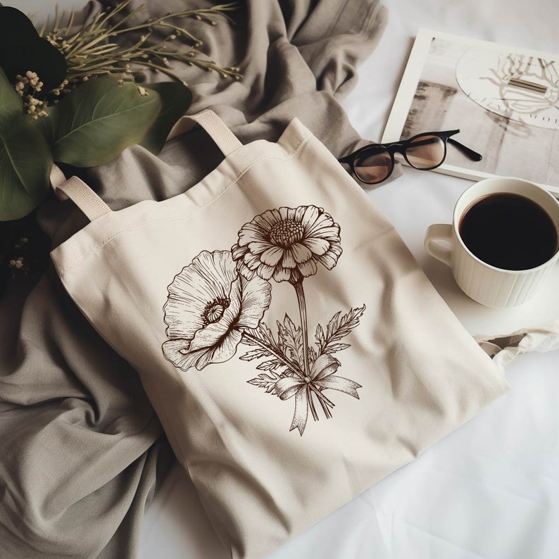 K&ouml;nnte beinhalten: Eine wei&szlig;e Canvas-Tragetasche mit braunen floralen Illustrationen von zwei Blumen, eine mit einer gro&szlig;en Mitte und die andere mit einer kleineren Mitte. Die Blumen sind mit einem Band zusammengebunden.