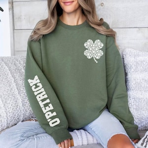 May include: An olive green sweatshirt featuring a white four-leaf clover design on the chest. The left sleeve displays the text "O'PETRICK" in white block letters. The sweatshirt is a crew neck style.