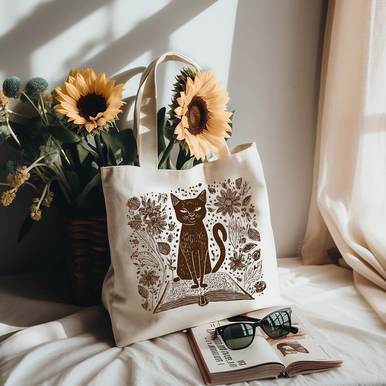 May include: A white canvas tote bag with a brown illustration of a cat sitting on a book surrounded by flowers. The cat has a serious expression and is looking directly at the viewer.