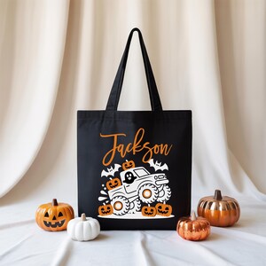 A black tote bag with a Halloween-themed design, including a truck and jack-o-lanterns, is displayed in the center of the image. Surrounding the bag are four small pumpkins, creating a festive and seasonal atmosphere.