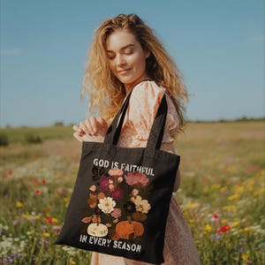 A young woman stands in a field of colorful wildflowers, holding a black tote bag with a floral design and text that reads "God is FAITHFUL in EVERY SEASON".