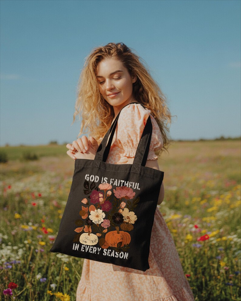 A young woman stands in a field of colorful wildflowers, holding a black tote bag with a floral design and text that reads "God is FAITHFUL in EVERY SEASON".