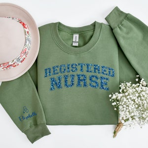 May include: A green sweatshirt with the text "REGISTERED NURSE" in blue glitter lettering. The sweatshirt has a small stethoscope and the name "Elizabeth" embroidered on the left sleeve.