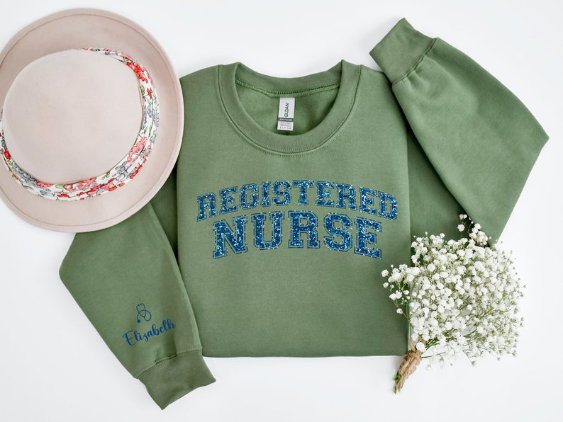 May include: A green sweatshirt with the text "REGISTERED NURSE" in blue glitter lettering. The sweatshirt has a small stethoscope and the name "Elizabeth" embroidered on the left sleeve.