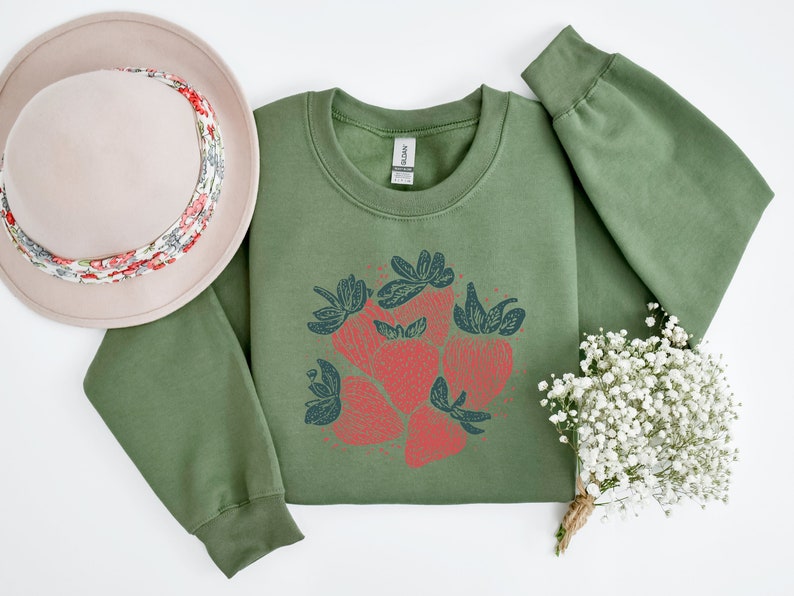May include: A green sweatshirt with a red and blue graphic of strawberries on the front. The sweatshirt is laid flat on a white surface with a hat and a bouquet of white flowers.