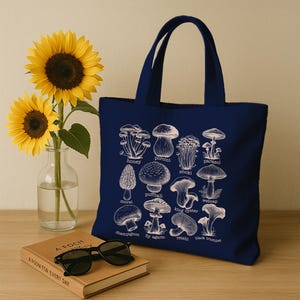 May include: A navy blue tote bag with a white illustration of different types of mushrooms. The mushrooms are labeled with their names, including honey, porcini, parasol, enoki, morel, puffball, king oyster, webcap, champignon, fly agaric, reishi, and black trumpet.