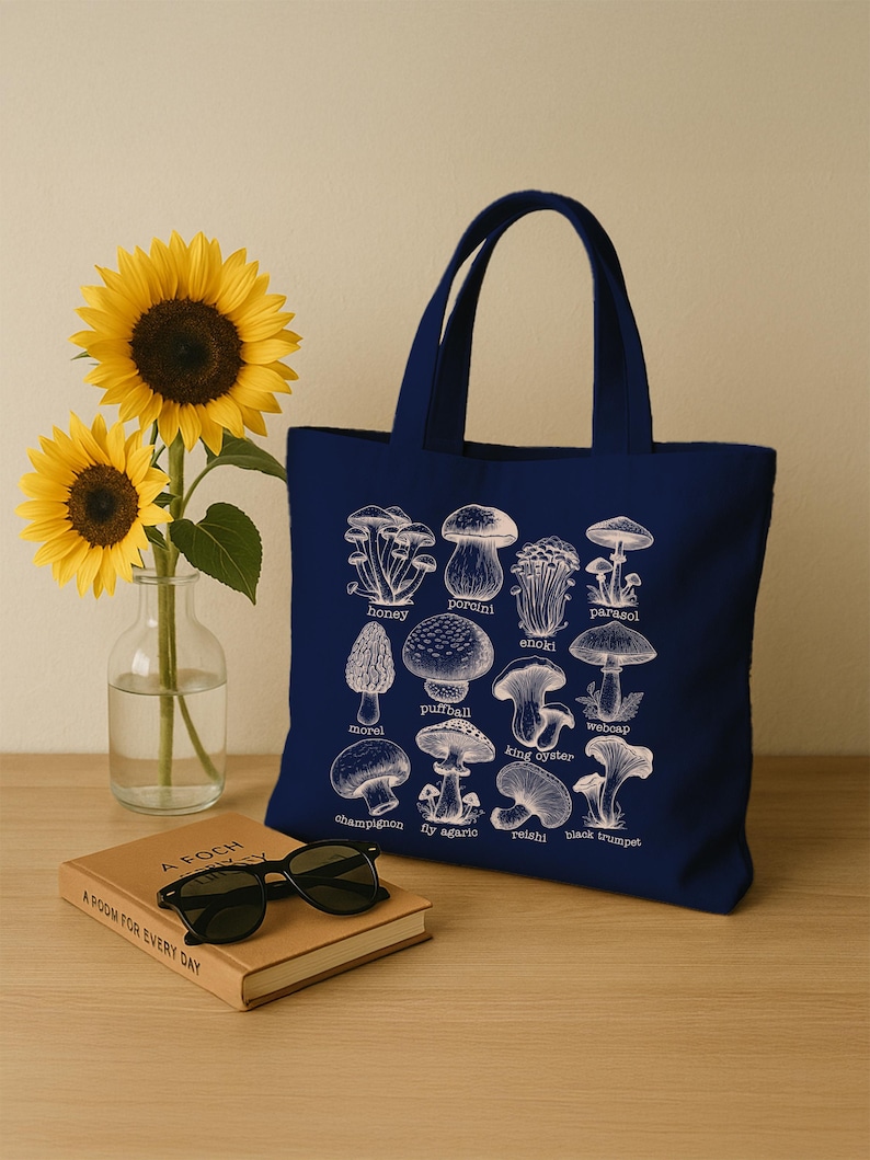 May include: A navy blue tote bag with a white illustration of different types of mushrooms. The mushrooms are labeled with their names, including honey, porcini, parasol, enoki, morel, puffball, king oyster, webcap, champignon, fly agaric, reishi, and black trumpet.