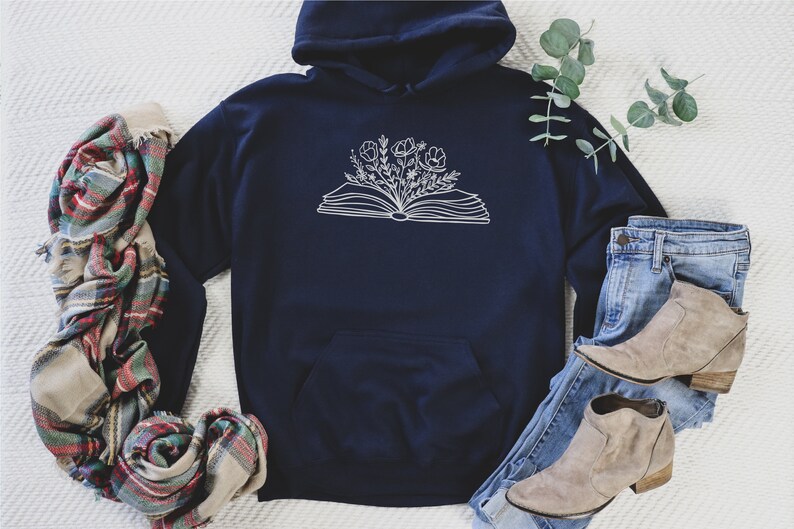 May include: A navy blue hoodie with a white outline of an open book with flowers growing from it. The hoodie is laying on a white surface with a plaid scarf and a pair of blue jeans and brown ankle boots.