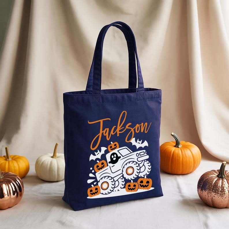 A navy blue tote bag with a Halloween-themed design, including a truck and jack-o-lanterns, is displayed in the center of the image. Surrounding the bag are small pumpkins, creating a festive atmosphere.