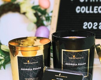 May include: Two candles with the text "Georgia Peach" on the label. One candle is gold, the other is black. The image also includes a small black sign that says "Spring Collection 2022". The background includes a wreath and Easter eggs.