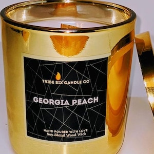 May include: A gold-colored candle in a glass jar with a matching lid. The candle has a black label with the text "Georgia Peach" and "Tribe Six Candle Co." The candle has a wood wick and is hand-poured.