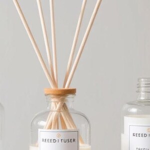 Puede incluir: Tres botellas difusoras de caña de vidrio transparente con etiquetas blancas y cañas de madera. La botella central tiene cañas insertadas en un collar de madera. Las etiquetas dicen "REED I FUSER".