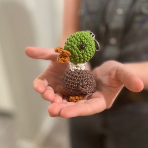 May include: A small crocheted duck keychain with green and brown yarn. The duck has a white beak and black eyes.