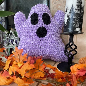 Könnte beinhalten: Ein flauschiges, lavendelfarbenes Geisterkissen mit schwarzen Augen und Mund. Der Geist ist von Herbstlaub und Halloween-Dekorationen umgeben, darunter ein Kerzenhalter in Form eines Spukhauses.