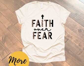 May include: A white t-shirt with a black cross and the text "FAITH over FEAR" in black letters with a leopard print background.