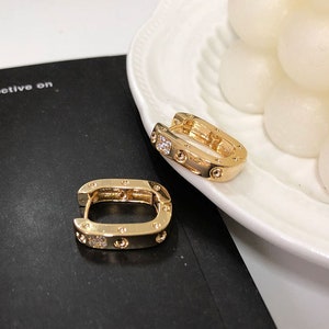 May include: Two gold hoop earrings with small clear stones set into the metal. The earrings are on a black surface.