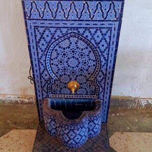 Puede incluir: Una fuente de azulejos azules y blancos con un arco decorativo y un grifo dorado. La fuente est&aacute; sobre un suelo de baldosas blancas. El texto "ceramiquefes" est&aacute; escrito en blanco sobre fondo negro.