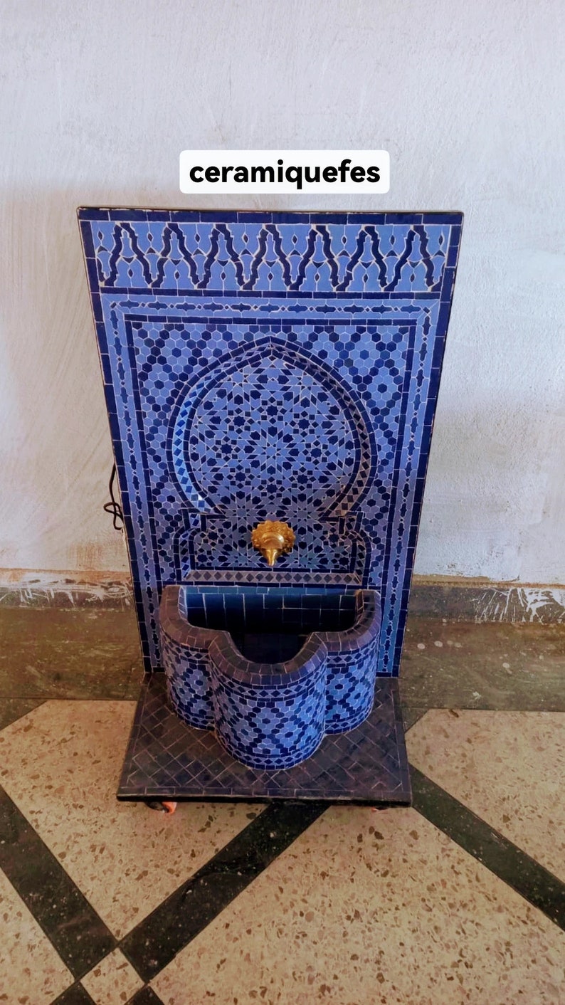 Puede incluir: Una fuente de azulejos azules y blancos con un arco decorativo y un grifo dorado. La fuente est&aacute; sobre un suelo de baldosas blancas. El texto "ceramiquefes" est&aacute; escrito en blanco sobre fondo negro.
