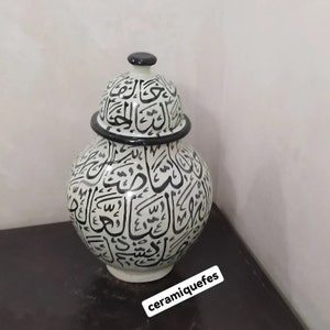 May include: White ceramic jar with a black lid, decorated with black Arabic calligraphy. The jar is sitting on a dark surface.