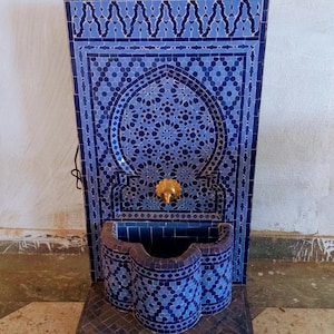 Puede incluir: Una fuente de azulejos azules y blancos con un grifo dorado. La fuente est&aacute; sobre ruedas y tiene un dise&ntilde;o decorativo. El texto "Ceramiqueefes" es visible sobre la fuente.