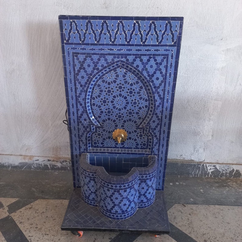 Puede incluir: Una fuente de azulejos azules y blancos con un arco decorativo y una peque&ntilde;a cuenca. La fuente est&aacute; sobre una base negra con ruedas.