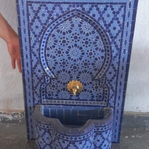 Puede incluir: Una fuente de azulejos azules y blancos con un arco decorativo y un ca&ntilde;o de lat&oacute;n. La fuente est&aacute; sobre una base negra con ruedas.