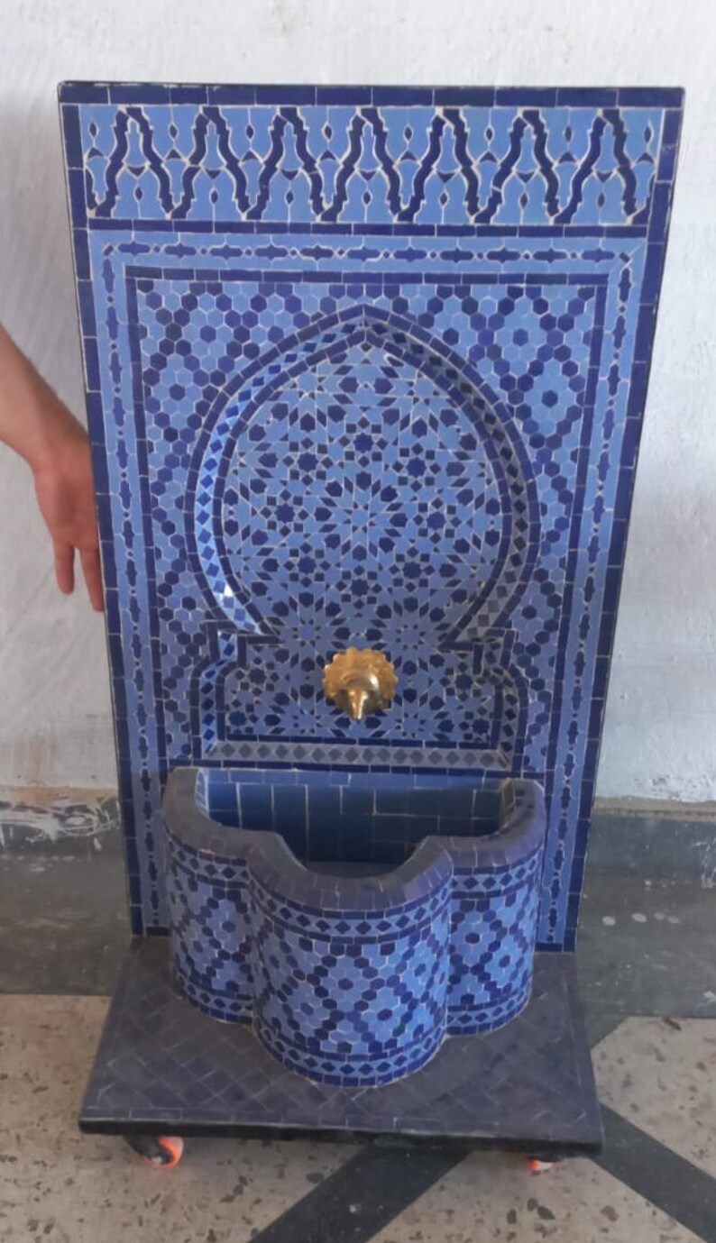 Puede incluir: Una fuente de azulejos azules y blancos con un arco decorativo y un ca&ntilde;o de lat&oacute;n. La fuente est&aacute; sobre una base negra con ruedas.