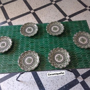 May include: Five green and white ceramic plates with intricate geometric designs on a green tiled table. The plates are all the same size and have a white background with green and black designs. The table is made of green tiles and has a black metal frame. The text "Ceramquefes" is visible in the bottom right corner of the image.