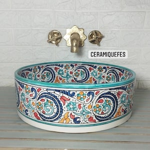 May include: A ceramic sink with a colourful floral design in blue, red, orange, and turquoise. The sink has a white base and a turquoise rim. The text "CERAMIQUEFES" is visible. Gold-coloured tap and fixtures are mounted on the wall.