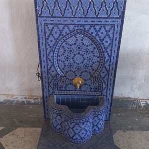 Puede incluir: Una fuente de azulejos azules y blancos con un arco decorativo y una peque&ntilde;a cuenca. La fuente est&aacute; sobre ruedas y tiene un grifo dorado.