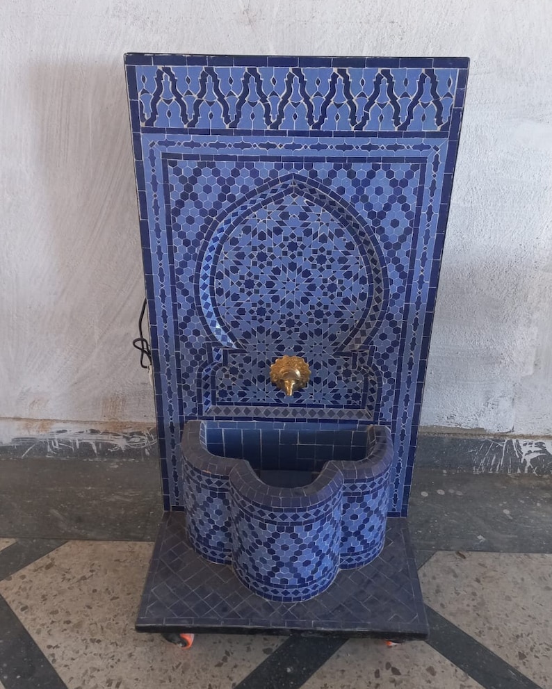 Puede incluir: Una fuente de azulejos azules y blancos con un arco decorativo y una peque&ntilde;a cuenca. La fuente est&aacute; sobre ruedas y tiene un grifo dorado.