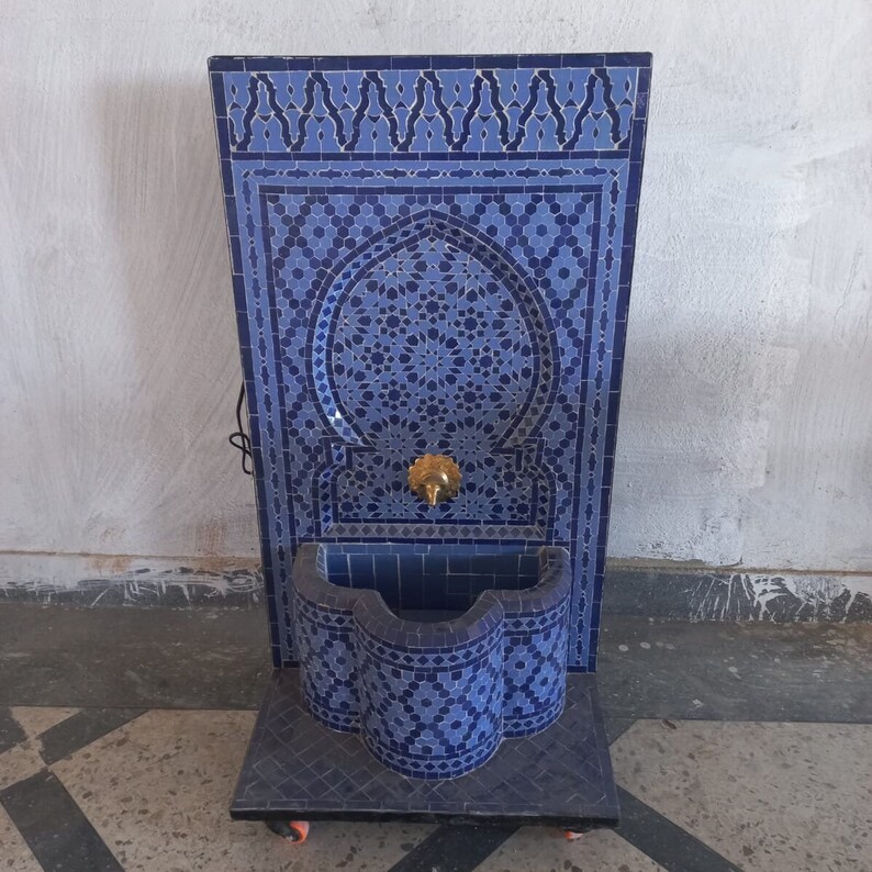 Puede incluir: Una fuente de azulejos azules y blancos con un arco decorativo y un ca&ntilde;o de lat&oacute;n. La fuente est&aacute; montada en un soporte con ruedas.