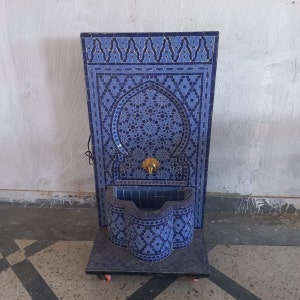 Puede incluir: Una fuente de azulejos azules y blancos con un arco decorativo y un grifo dorado. La fuente est&aacute; sobre una base negra con ruedas.
