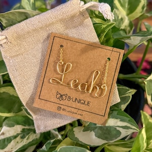 Puede incluir: Collar de alambre dorado con el nombre "Leah" escrito en cursiva. El collar se muestra en una tarjeta marrón con el texto "B-UNIQUE" y "@b_unique_usa".