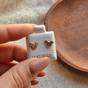 以下が含まれることがあります： 白いカードに展示された、金色のマウスヘッド型のスタッドピアス。カードには「10 KT. GOLD」の文字が金色で印刷されています。ピアスは小さくて光沢があります。